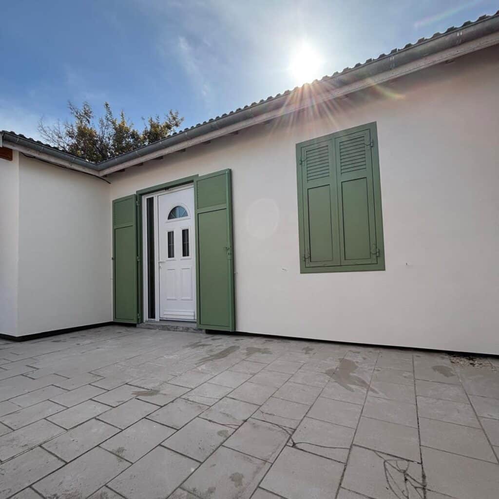 Façade de Maison avec Volets Verts et Terrasse Extérieure Façade de maison blanche, volets battants verts, porte d'entrée blanche et terrasse carrelée sous le soleil.